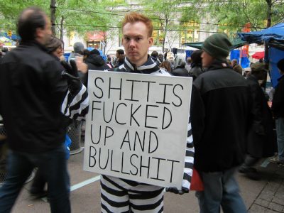 Protestor at Occupy Wall Street dressed in a black and white striped suit ala prison garb holding a hand lettered sign reading Shit is fucked up and bullshit. Photo by Scott Lynch